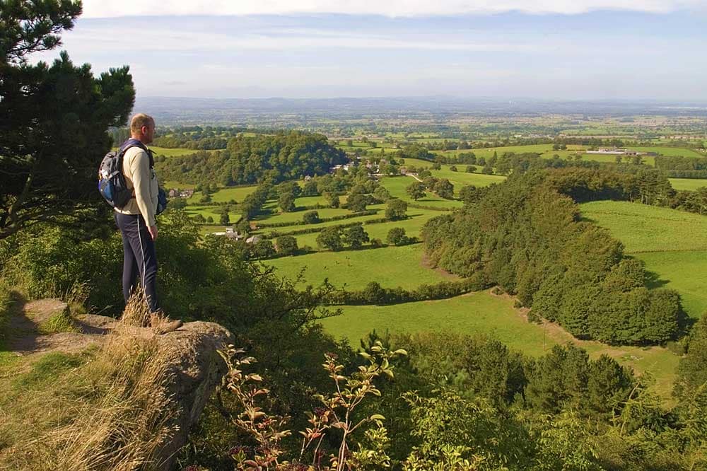 Hiker standing on a rocky outcrop overlooking the lush green countryside of the Sandstone Ridge, Cheshire, UK.