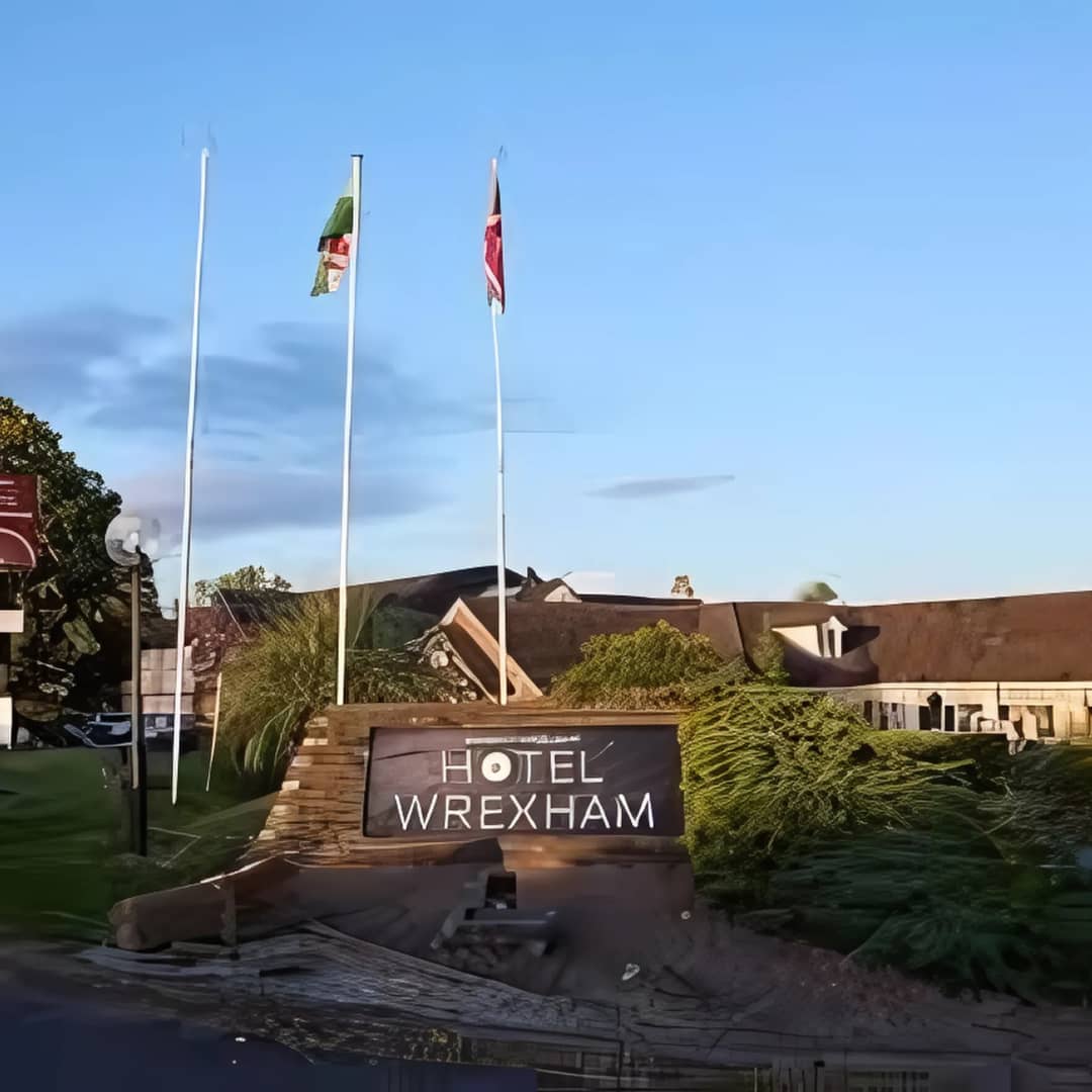 Hotel Wrexham entrance sign with two flagpoles flying the Welsh and Union Jack flags, set against a clear blue sky, surrounded by greenery and modern architecture.