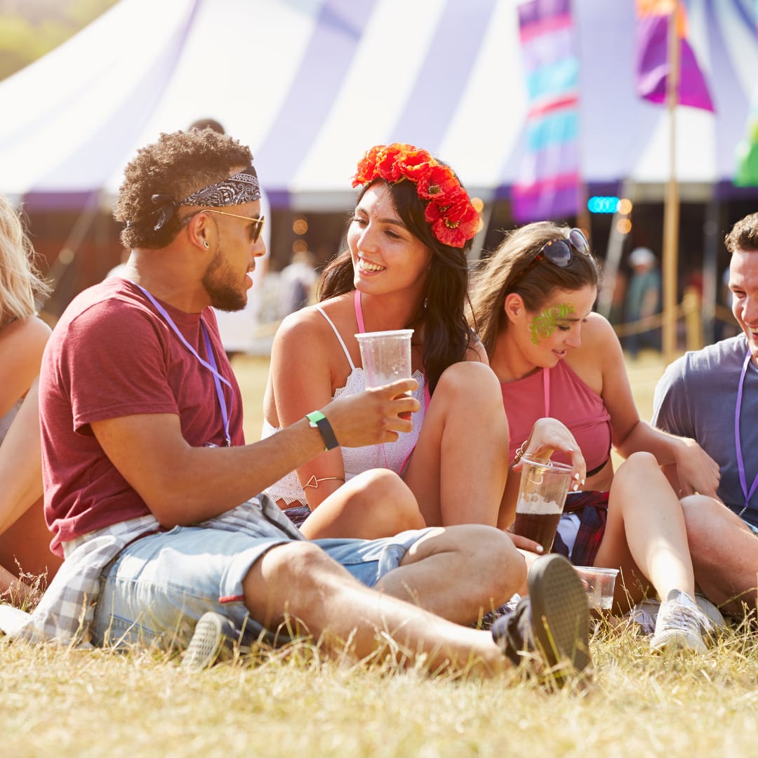 A group of young festival-goers relaxes on the grass at Together Again Festival, enjoying drinks, laughter, and good vibes under the summer sun, with colourful tents and flags in the background.