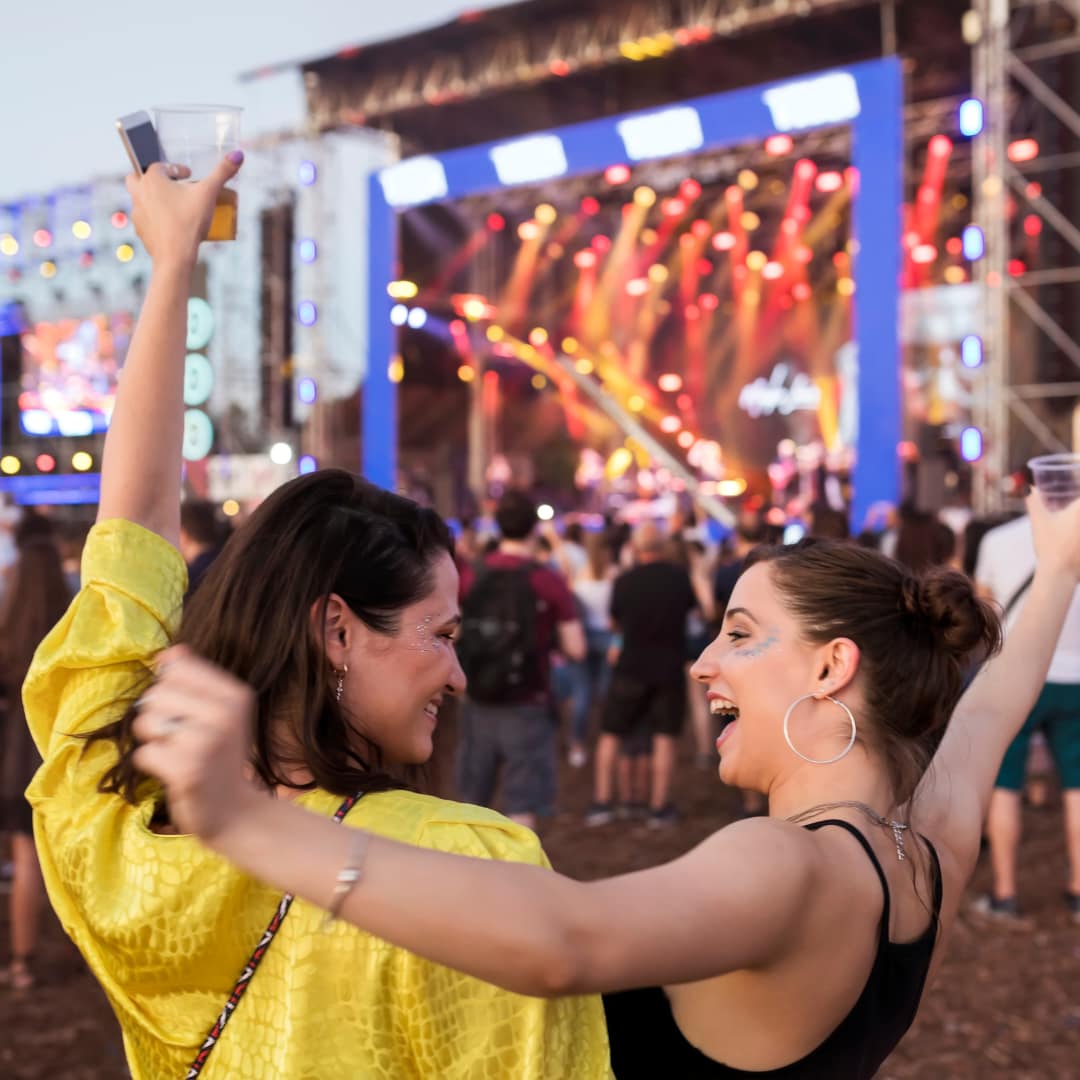 Two women celebrating at a live music festival, raising their hands in excitement while enjoying the vibrant atmosphere. A large stage with bright lights and a crowd of festivalgoers can be seen in the background.