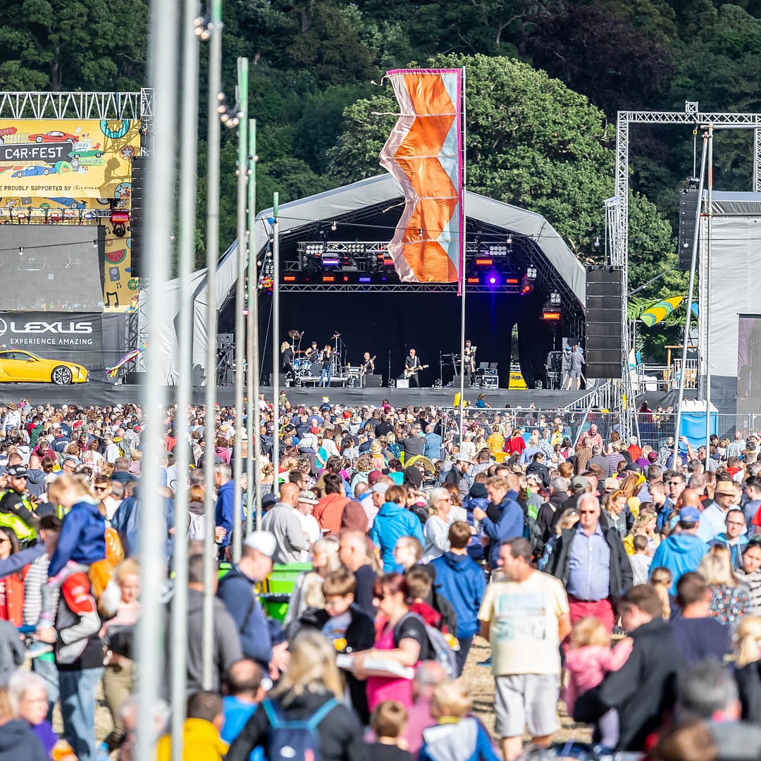 Large crowd gathered at the main stage of CarFest with colourful flags and a festival atmosphere.