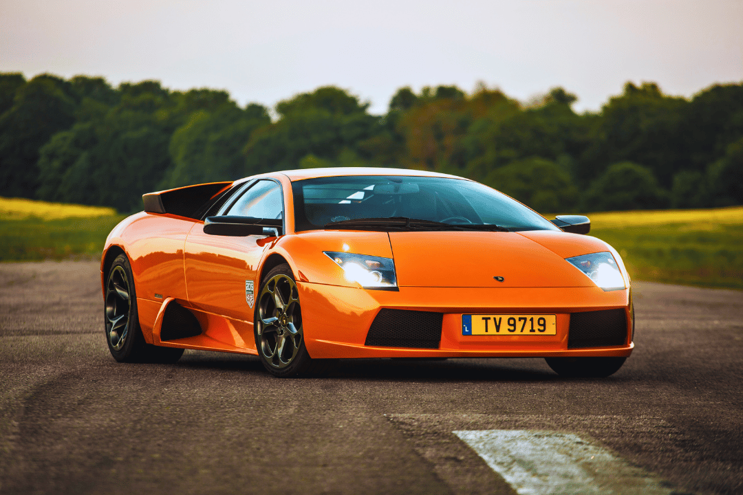 An orange sports car parked in the centre of a race track, with large trees in the background.