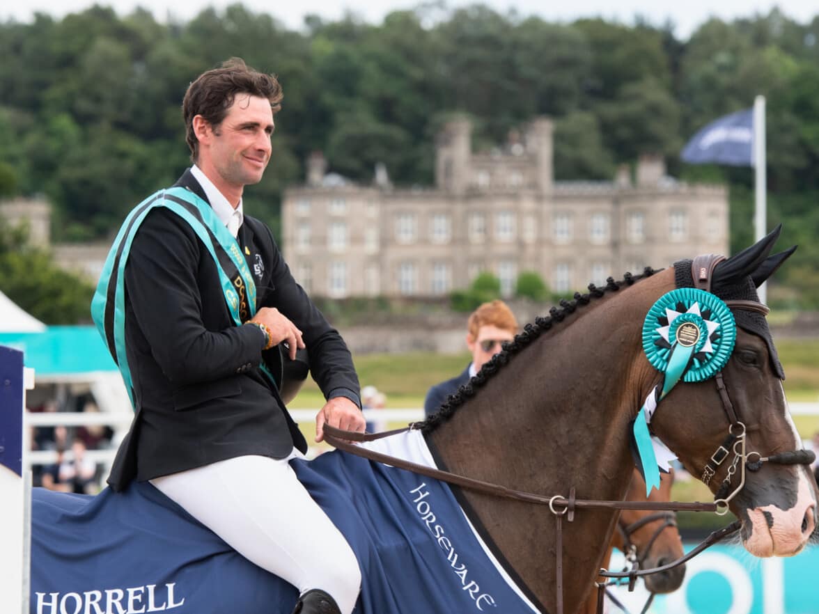 An equestrian winner riding a horse, wearing a champion sash and rosette, with Bolesworth Castle in the background