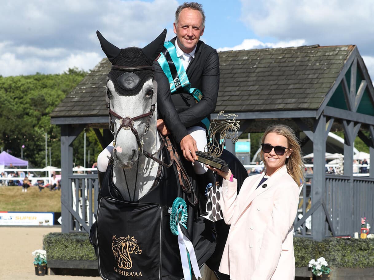 Award ceremony featuring a rider on a horse adorned in Al Shira'aa branding, accompanied by an official and a spectator near a presentation area.