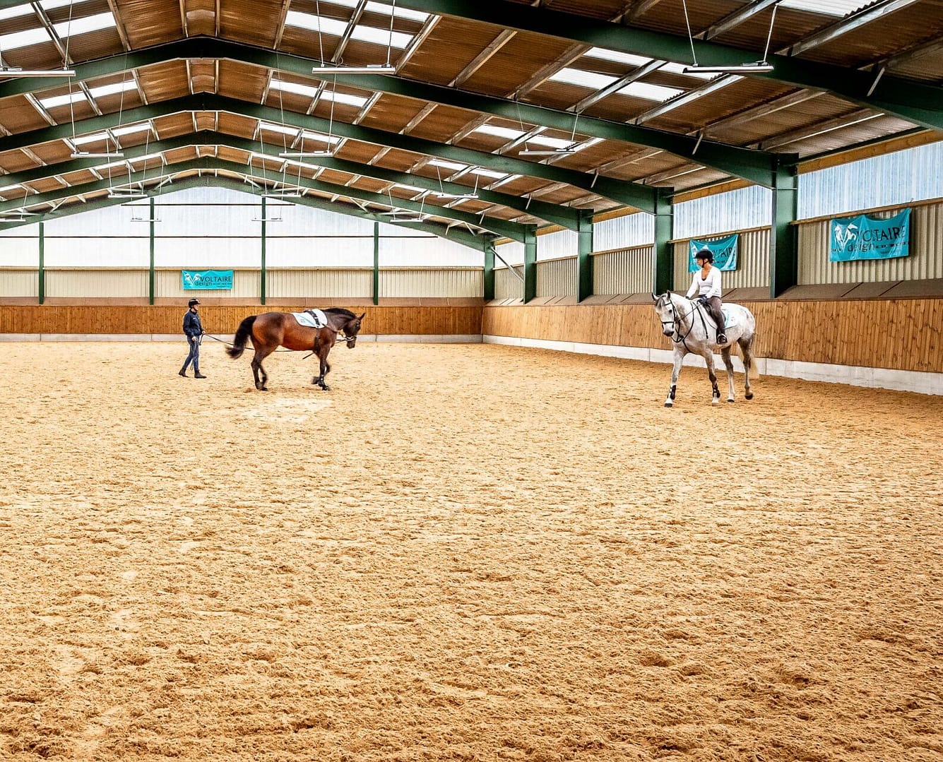 Indoor equestrian centre with two horses and riders.