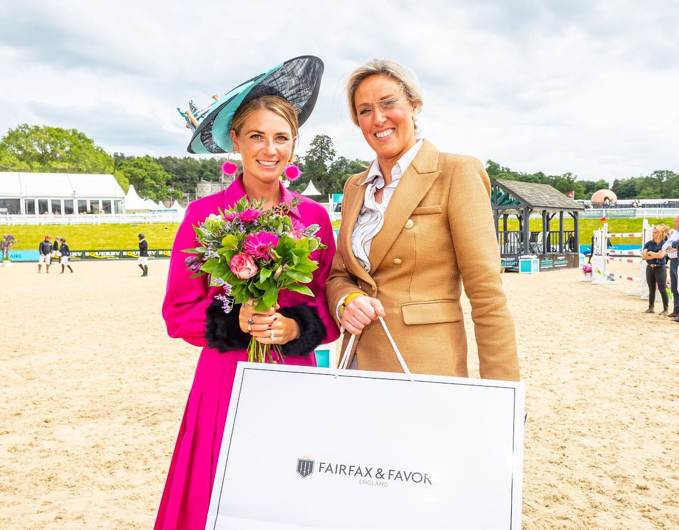 Two women pose at Bolesworth International, one holding a bouquet of pink and green flowers and the other carrying a Fairfax & Favor.