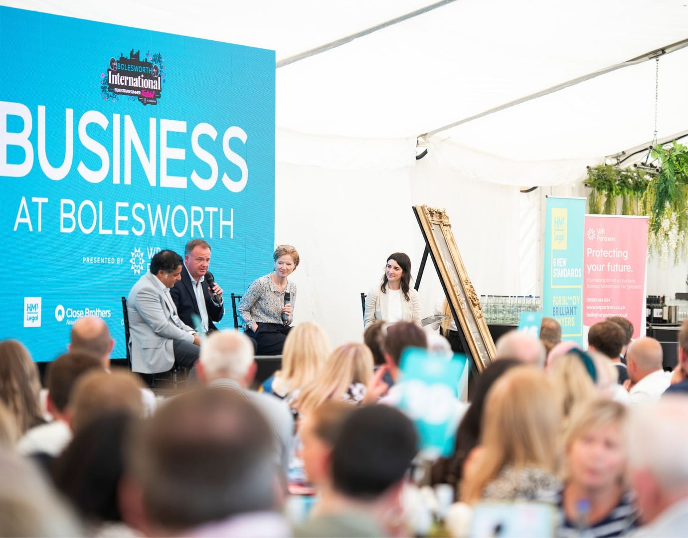 A lively business panel discussion at Bolesworth International, featuring three speakers seated on stage with microphones. A vibrant audience listens attentively, with the backdrop displaying the "Business at Bolesworth" branding.