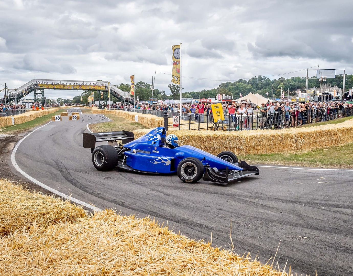 A blue race car with flame decals navigating a corner of a motorsport track surrounded by straw bales, with a crowd of spectators and a footbridge in the background.