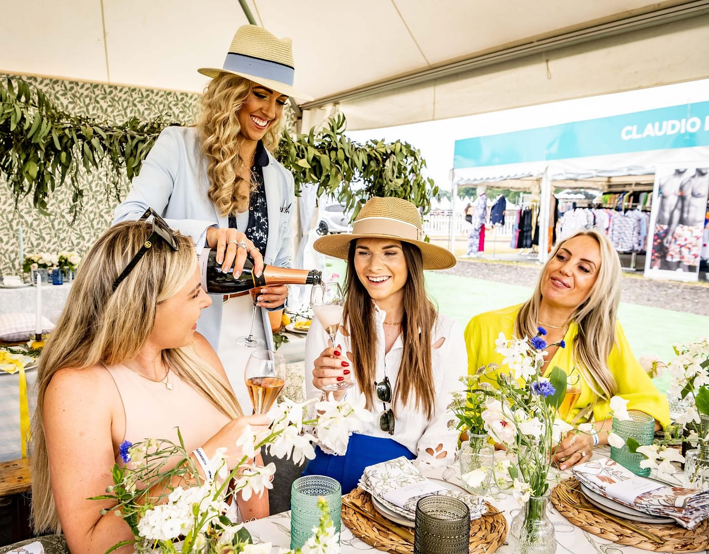 Four women enjoying a beautifully set table with floral arrangements, as one of them pours rosé wine into glasses under a marquee at Bolesworth.