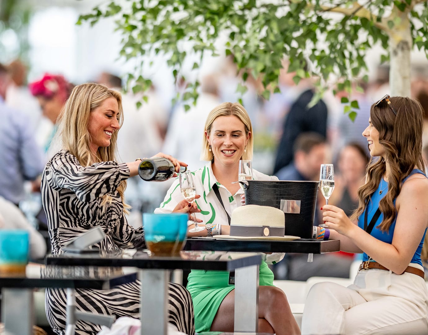 Three women seated at a table under a leafy tree, enjoying champagne and smiling in a lively event setting.