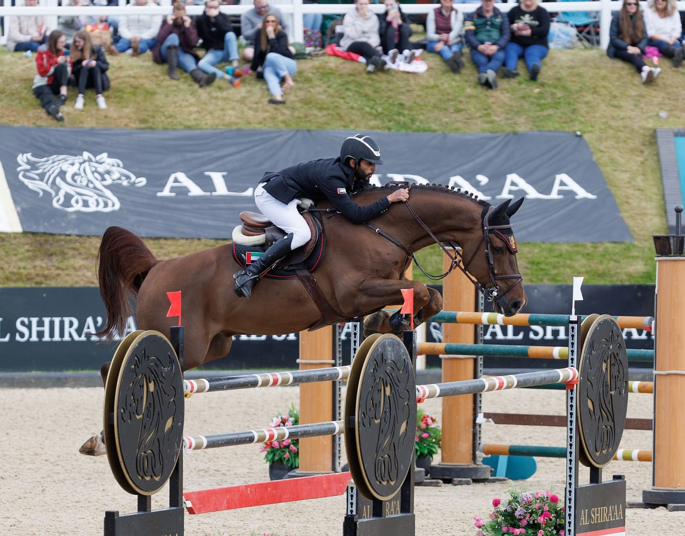 Al Shira'aa showjumping rider clears a jump at Bolesworth International Horse Show, with a focused crowd watching in the background.