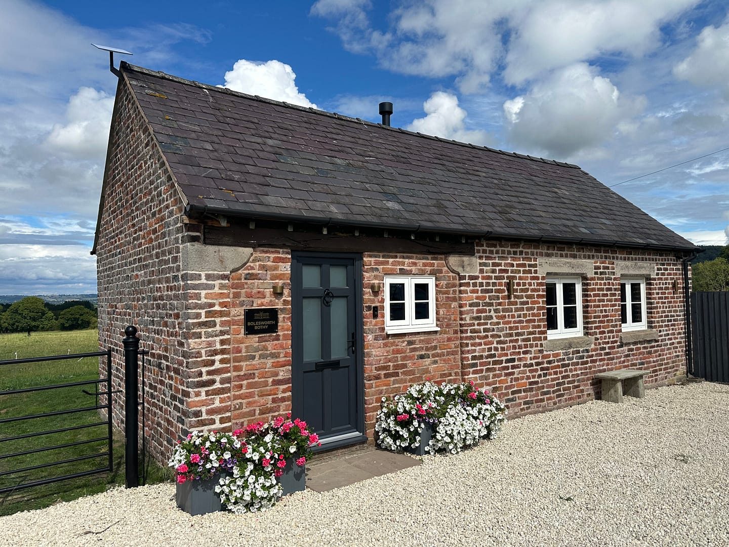 Small detached ground level house with planters with fresh flowers outside the front door on each side and a small bench underneath the window.