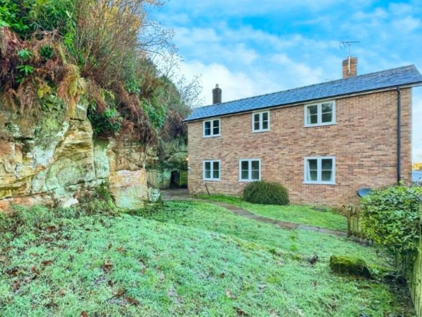 A modern brick-built detached house nestled against a natural rock face, featuring large windows, a well-maintained lawn, and a rural backdrop.