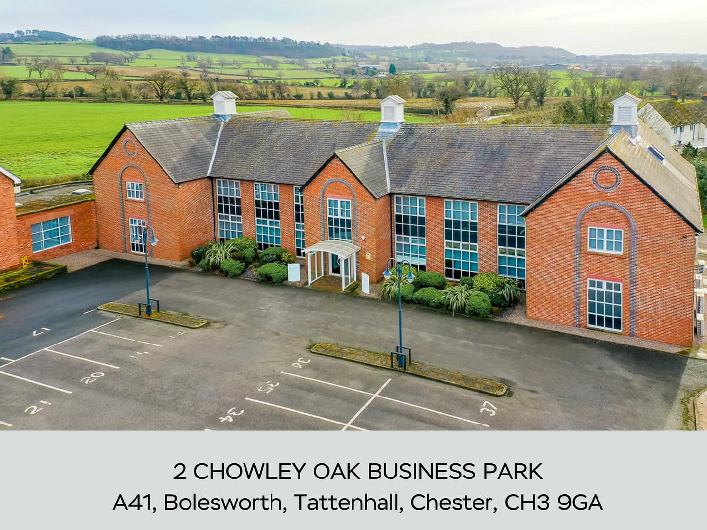 A modern red-brick office building with large glass windows, ample parking spaces, and landscaped greenery at 2 Chowley Oak Business Park, Bolesworth, Tattenhall, Chester.