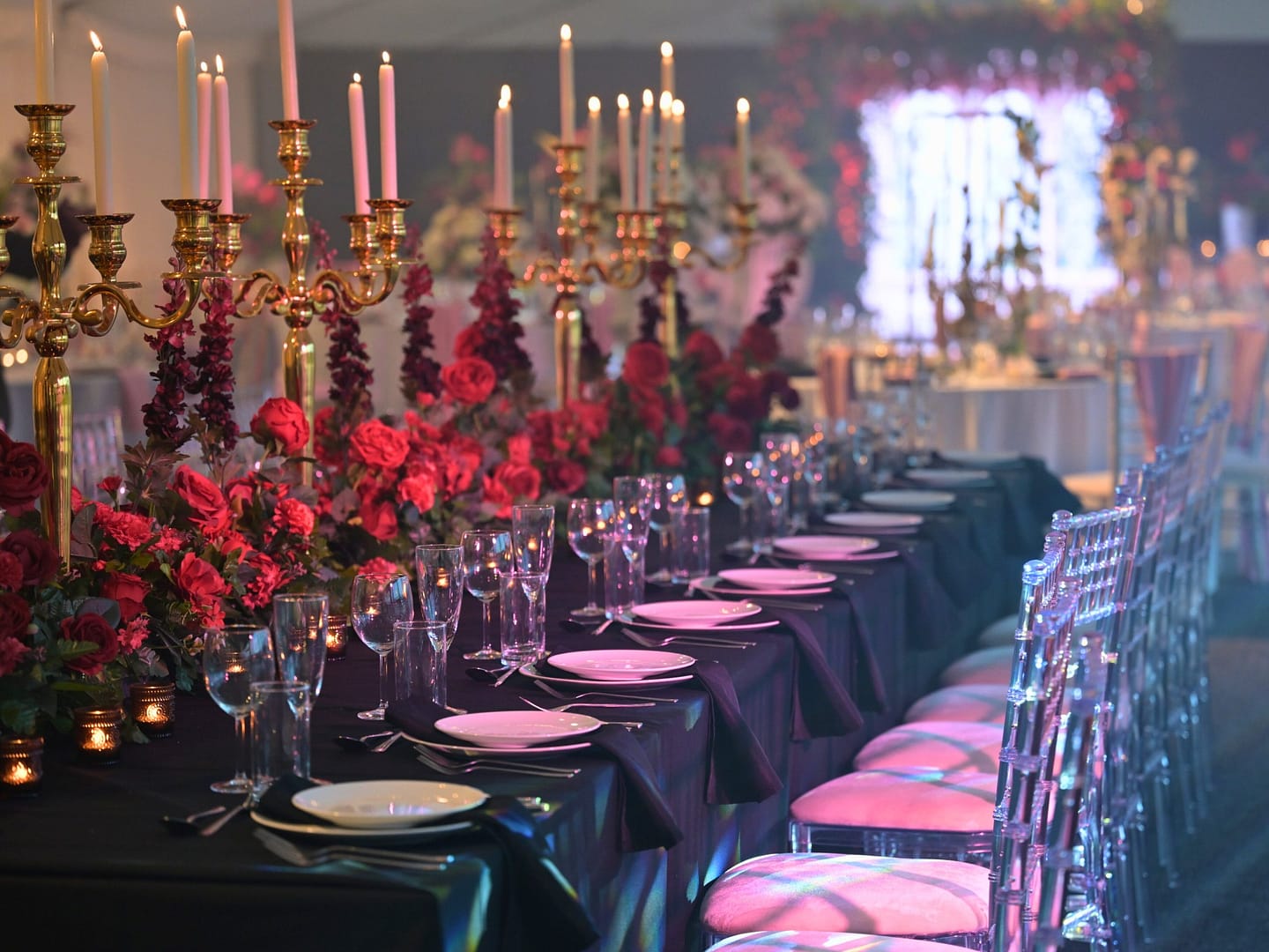 A beautifully arranged candlelit wedding reception table, featuring gold candelabras, rich floral arrangements with deep red roses, elegant table settings with black napkins, and crystal-clear glassware, creating a romantic and luxurious ambiance.