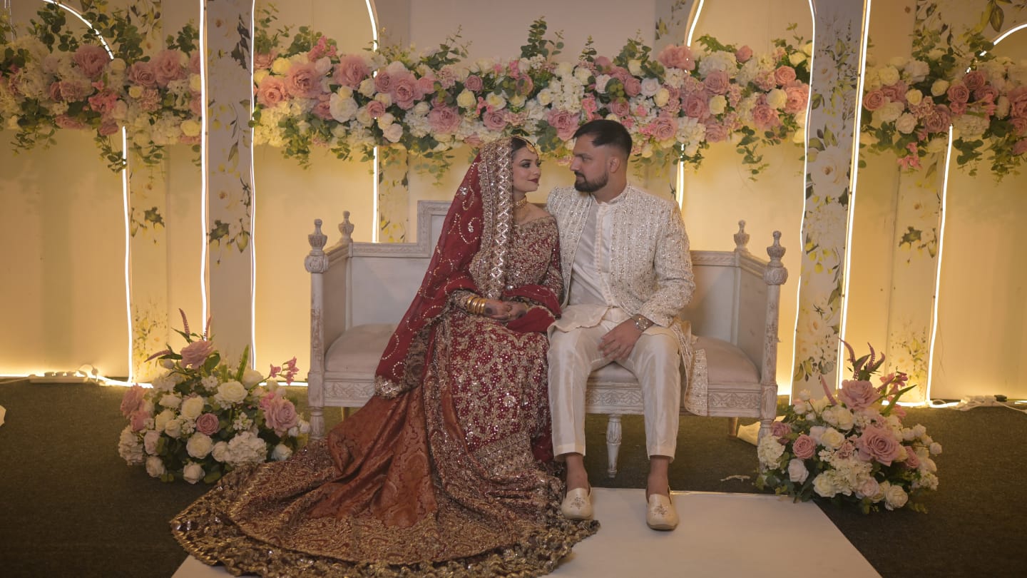 A South Asian wedding couple seated on an ornate ivory stage with floral arrangements and ambient lighting. The bride is wearing a traditional deep red and gold embellished bridal lehenga with intricate embroidery, jewelry, and a matching dupatta, while the groom dons an ivory sherwani with delicate gold accents. The romantic setting features soft pastel roses, white florals, and warm lighting, creating an enchanting wedding ambiance.