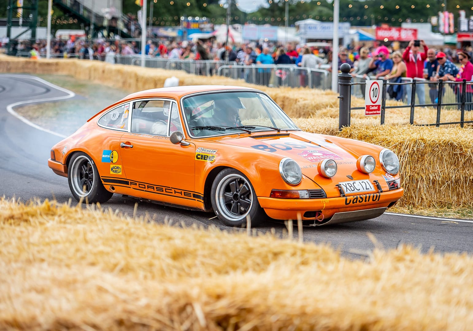 An small orange motor car racing around an internal track at Bolesworth during Carfest North with crowds watching and cheering on