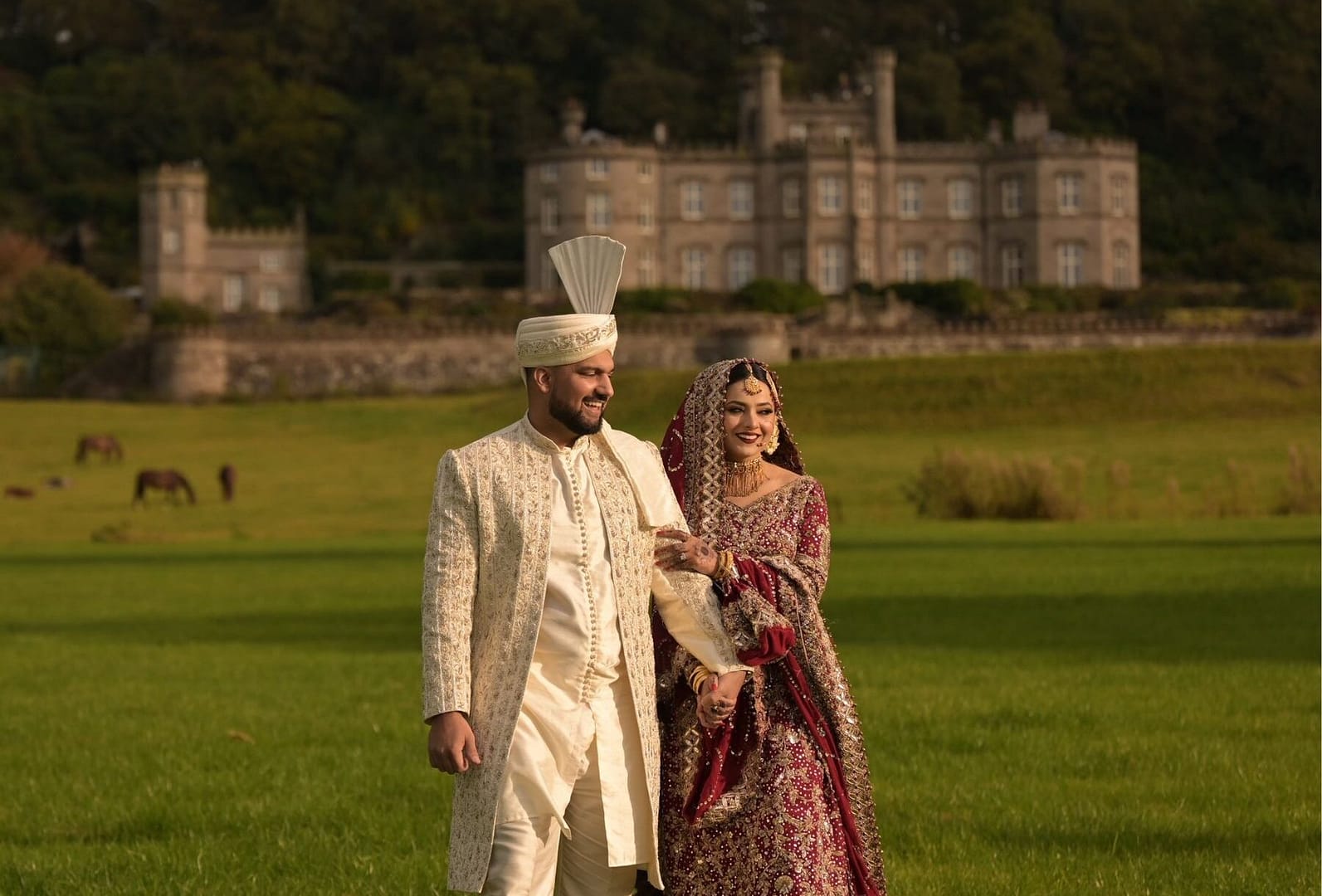 Elegant couple in traditional wedding attire walking on a lush green lawn with a historic estate in the background.
