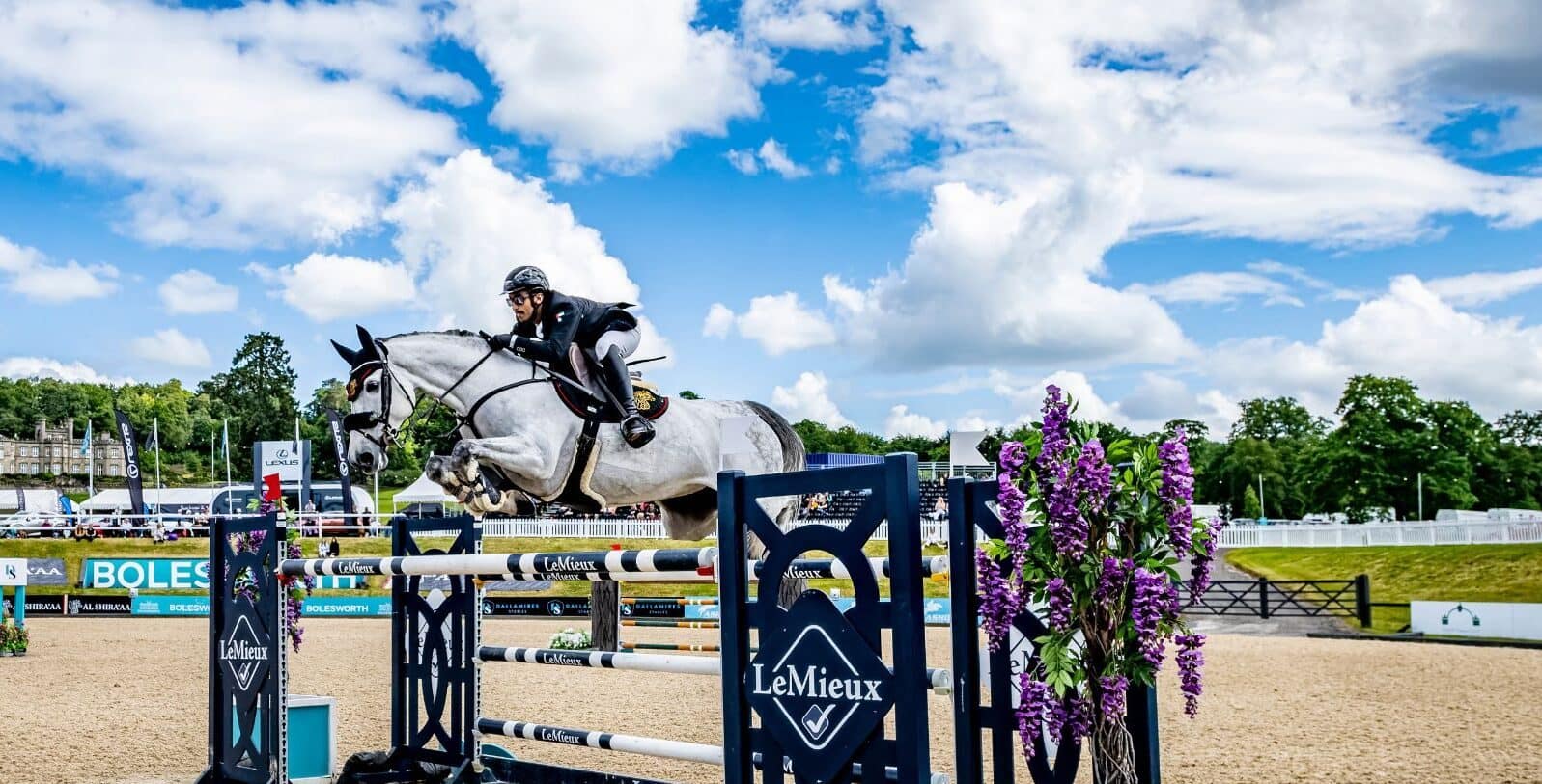 An equestrian rider expertly jumps over a decorative fence featuring "LeMieux" branding at the Bolesworth International. The backdrop showcases the Bolesworth estate, with vibrant trade stands.