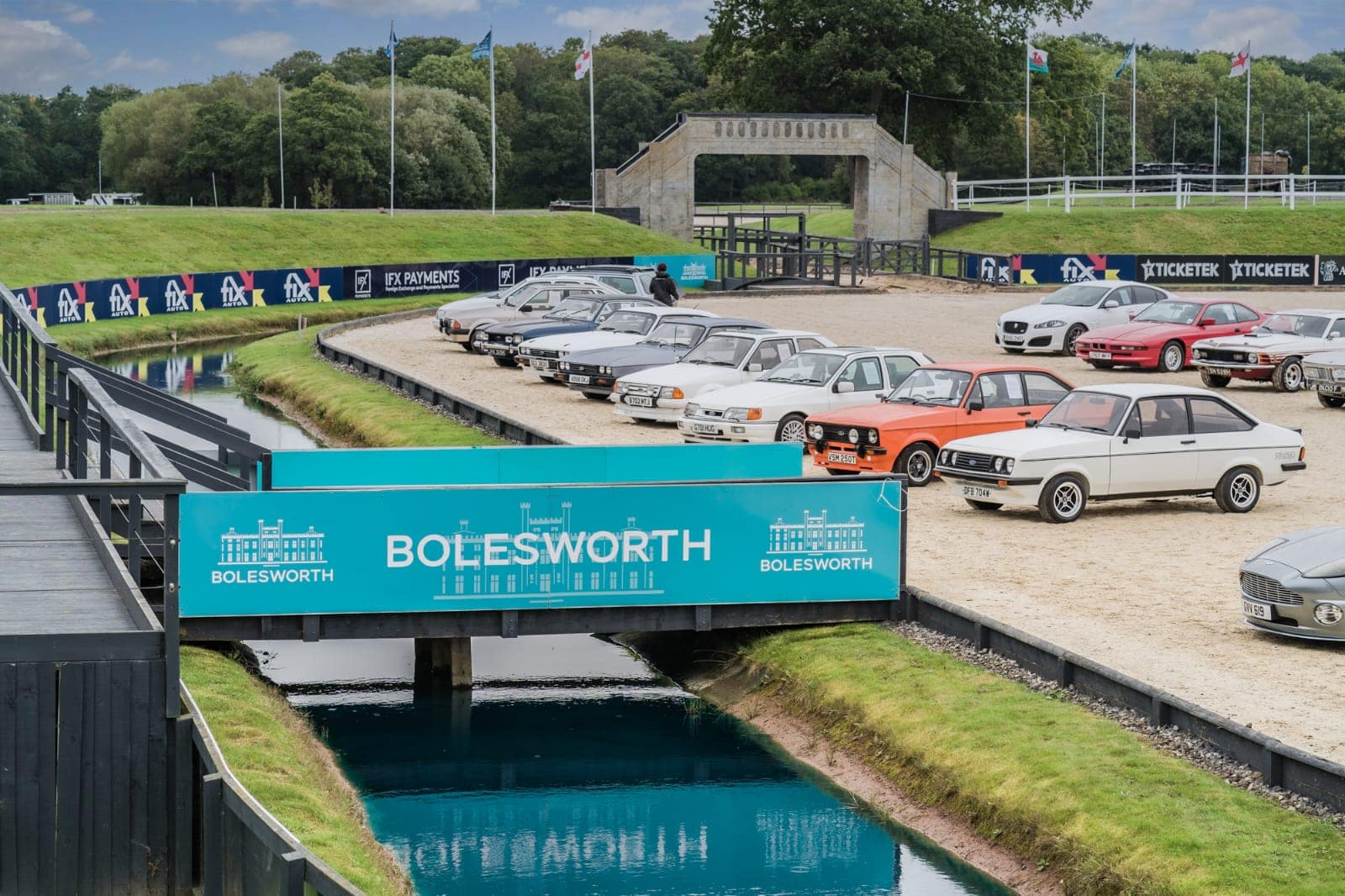 A collection of classic and vintage cars displayed at the Hampson Car Auction at Bolesworth Estate, Cheshire. The image features a variety of iconic Ford, Jaguar, and Aston Martin models, neatly arranged in a prestigious auction setting with Bolesworth branding on the bridge over a reflective waterway.