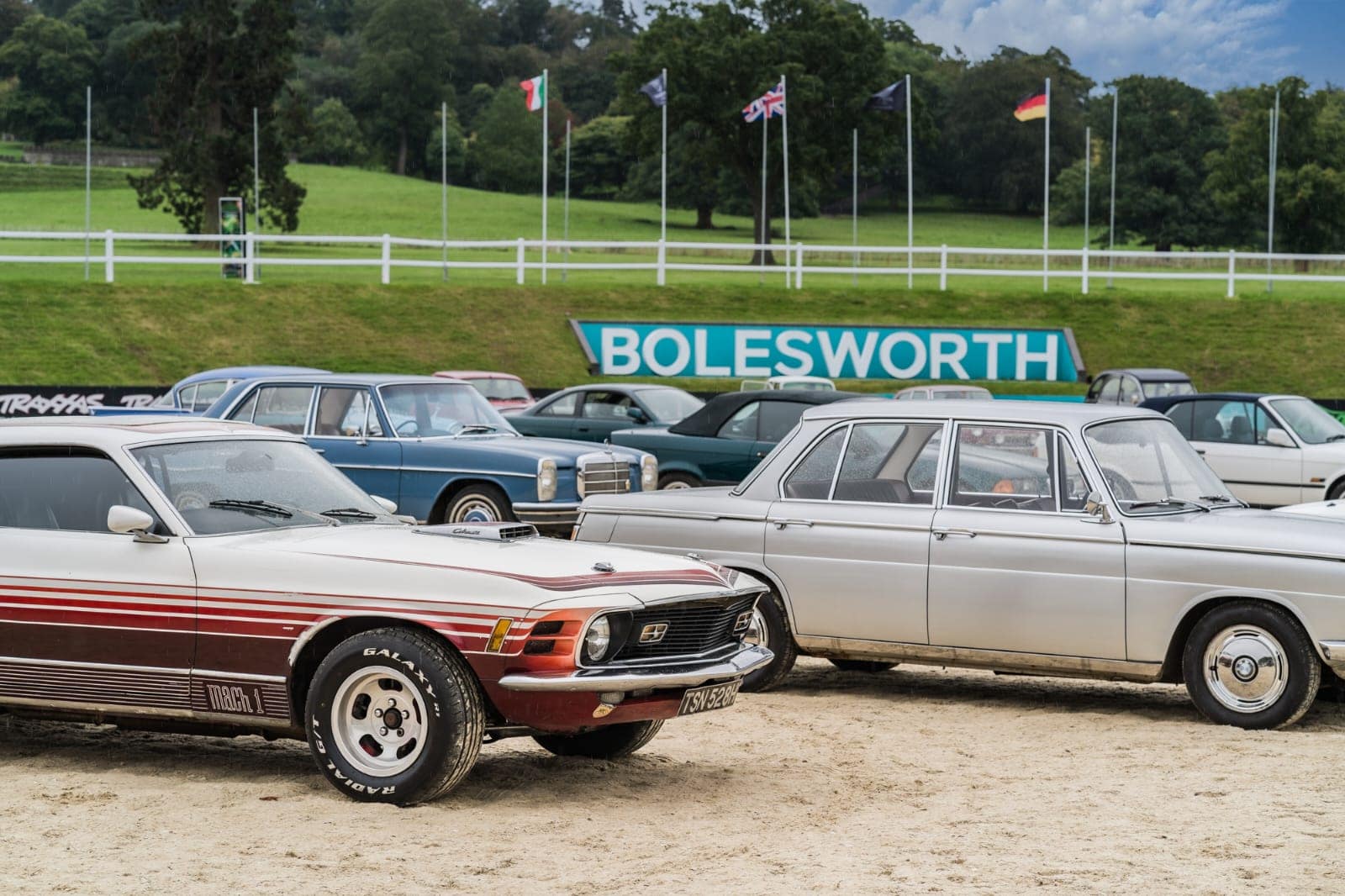 A 1970 Ford Mustang Mach 1 with racing stripes, a Mercedes-Benz classic saloon, and other vintage cars at the Hampson Car Auction at Bolesworth Estate, Cheshire. The backdrop features the Bolesworth sign, lush green hills, and international flags, enhancing the prestigious auction atmosphere.