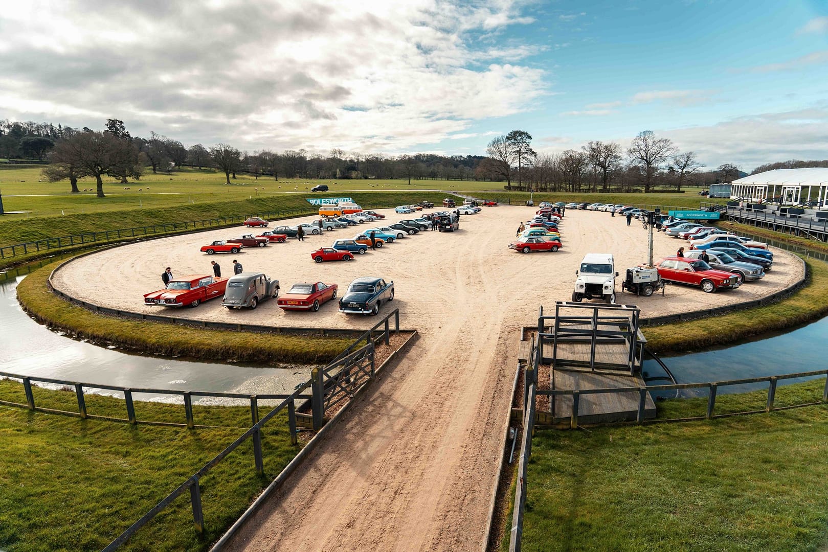 A stunning aerial view of the Hampson Classic Car Auction at Bolesworth, featuring a variety of vintage and luxury cars displayed in a circular sandy arena. Classic red sports cars, vintage saloons, and 4x4 vehicles are showcased, surrounded by green countryside under a partly cloudy sky.