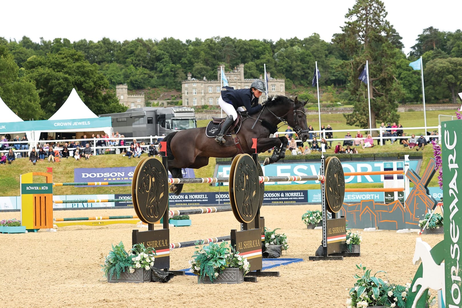 Louise Saywell and Kingsborough Kasper clearing a showjumping obstacle at Bolesworth International Horse Show, with a crowd and Bolesworth Castle in the background.