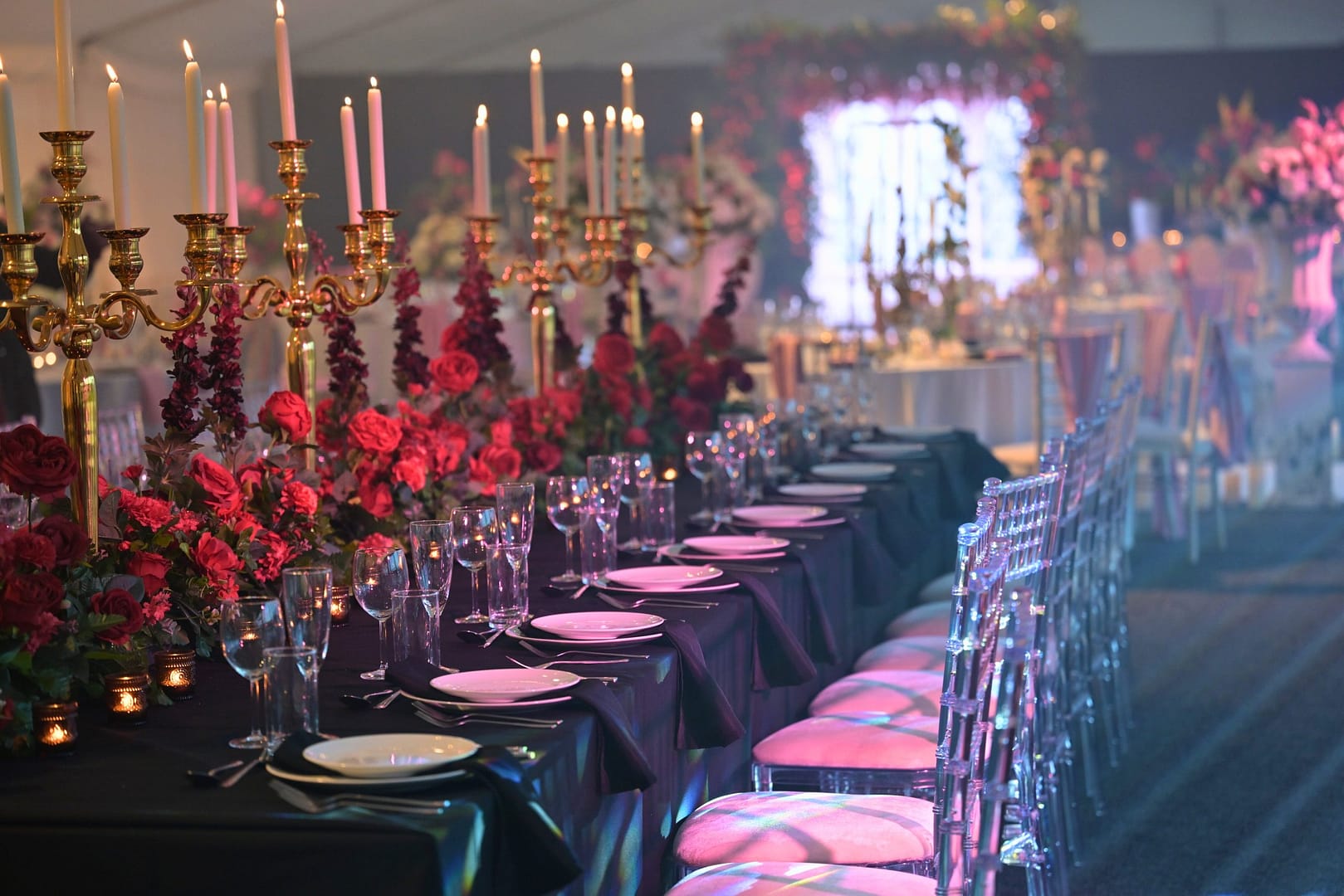 A beautifully arranged candlelit wedding reception table, featuring gold candelabras, rich floral arrangements with deep red roses, elegant table settings with black napkins, and crystal-clear glassware, creating a romantic and luxurious ambiance.