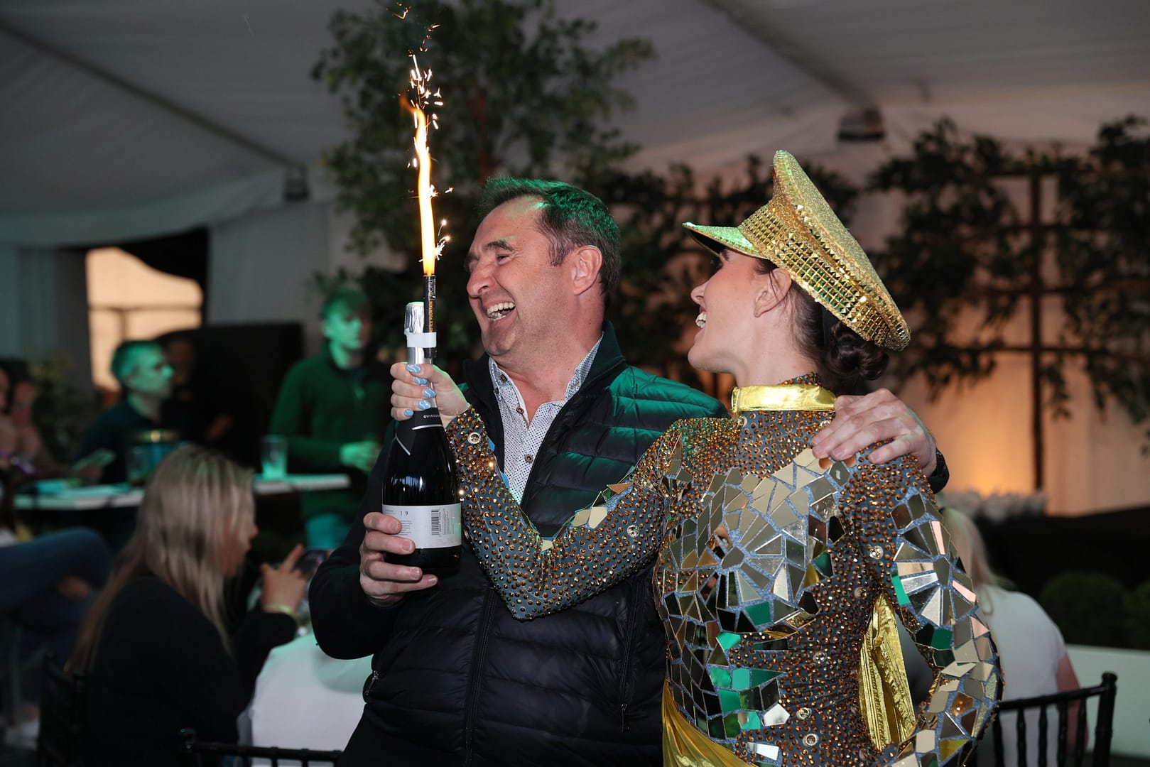 A woman wearing a shiny gold diamanté theatre hat holds a bottle of champagne with a lit sparkler attached, standing next to a well-dressed gentleman in a puffer jacket and shirt.