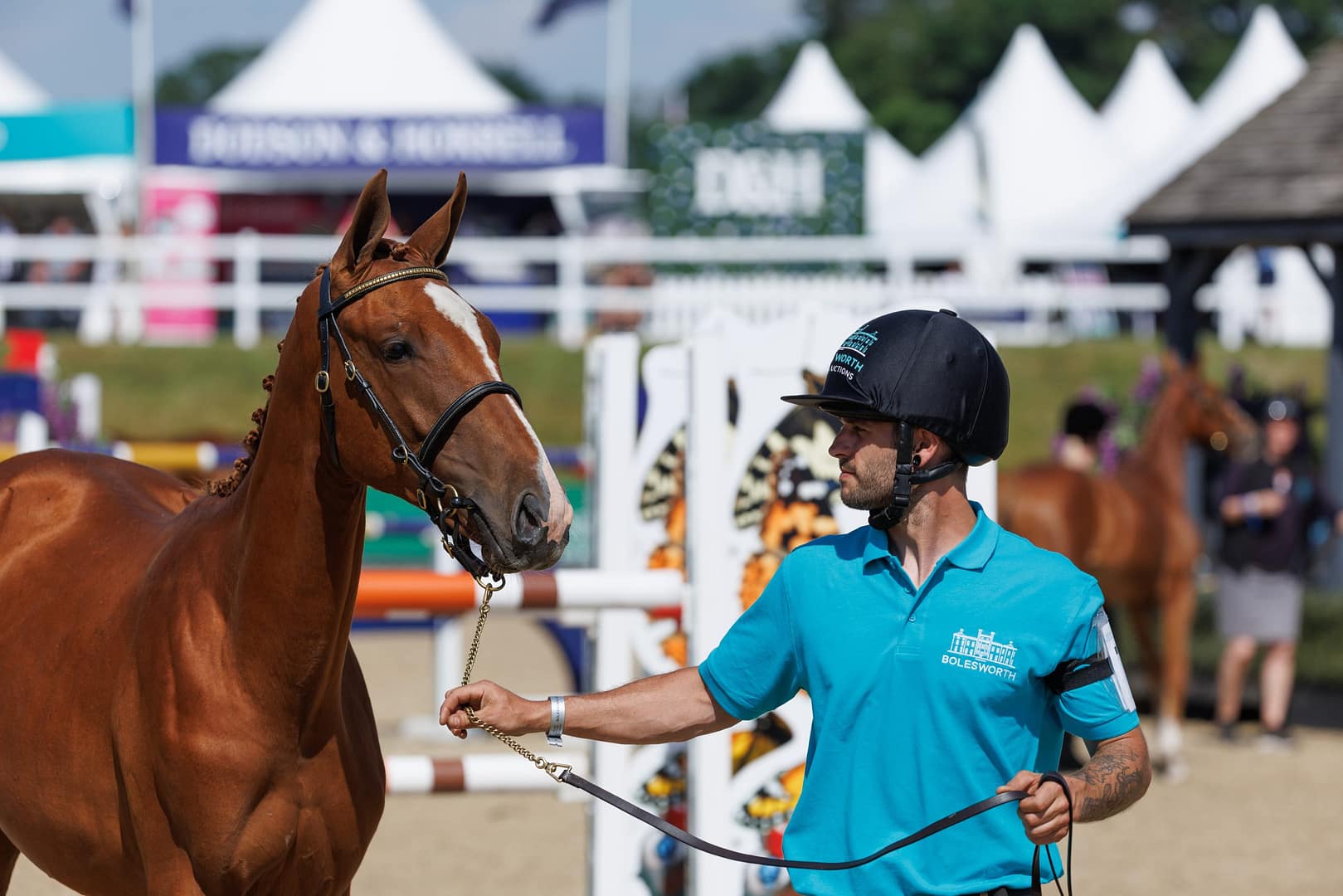 A chestnut foal with a white blaze is presented by a handler wearing a Bolesworth-branded polo shirt and helmet at the Bolesworth International Horse Show 2023. The foal stands alert in front of a showjumping arena with luxury tents and sponsor banners in the background.