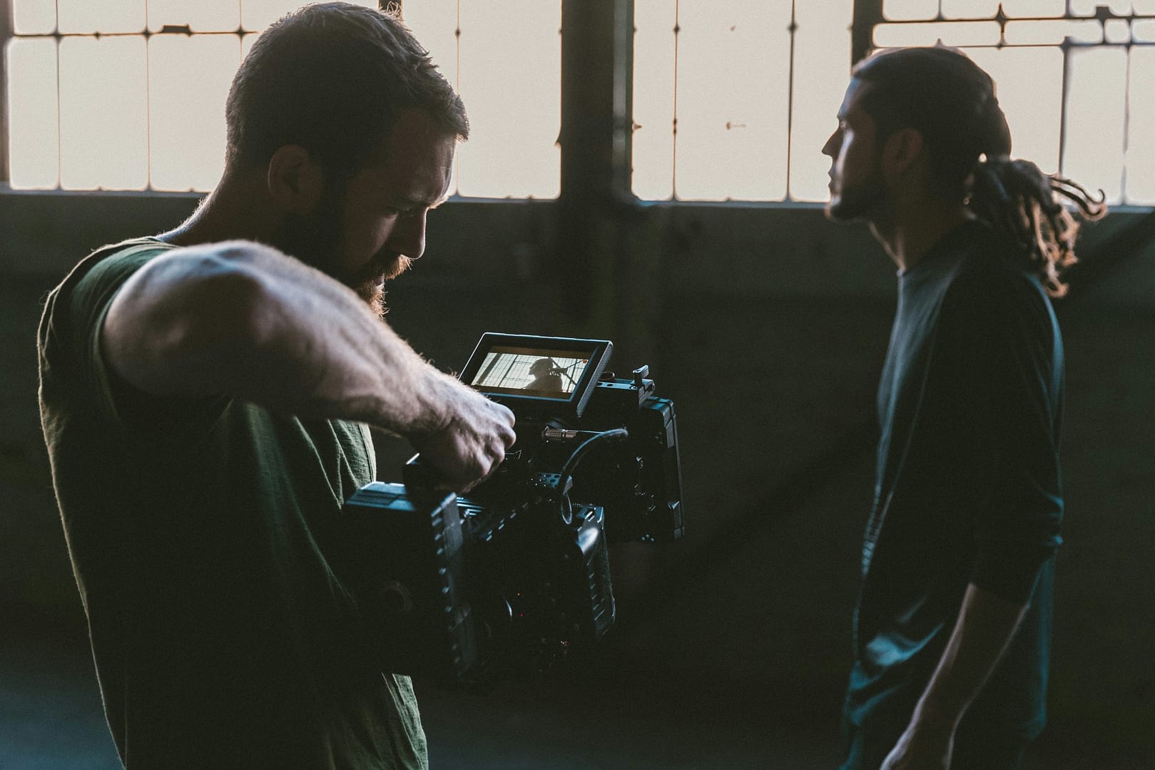 A filmmaker operates a professional cinema camera, capturing a dramatic scene with a silhouetted actor in a dimly lit industrial setting. The focus is on high-end film production, lighting, and storytelling.