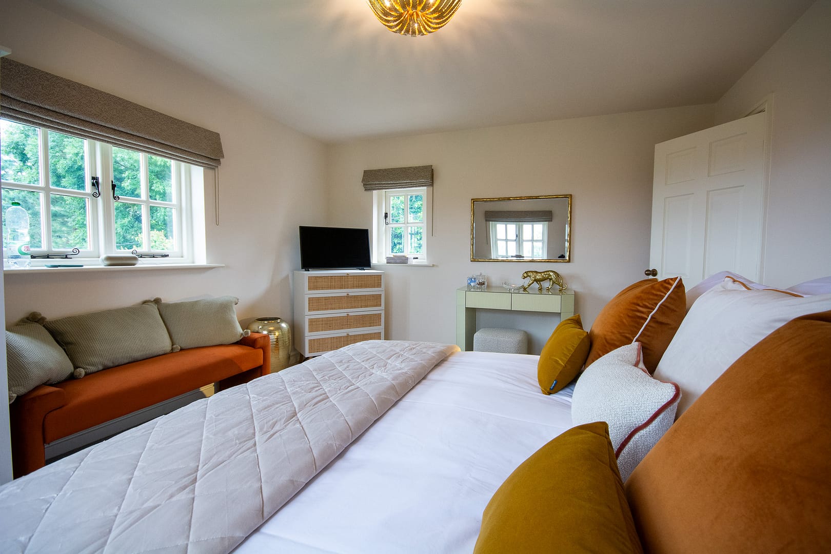 A bedroom with two windows. The bed has white linen and orange and mustard scatter cushions. Underneath the window is an orange bench