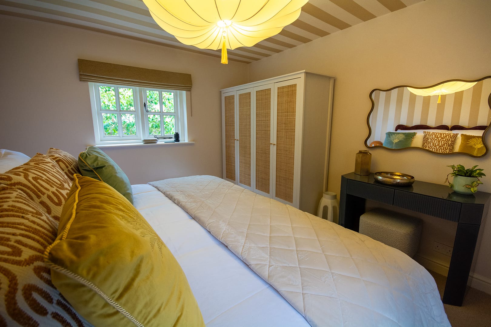 A bedroom with large bed with scatter cushions, free standing wardrobe, desk and mirror and a window