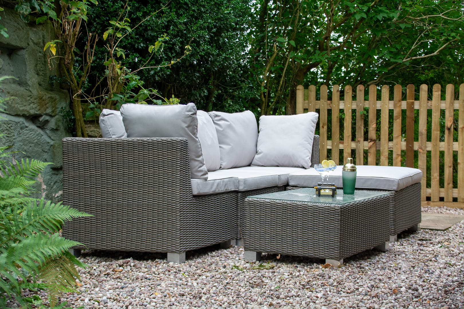 Grey rattan sofa with scatter cushions and low coffee table sitting in a secluded area with a small garden fence and pebble on the ground