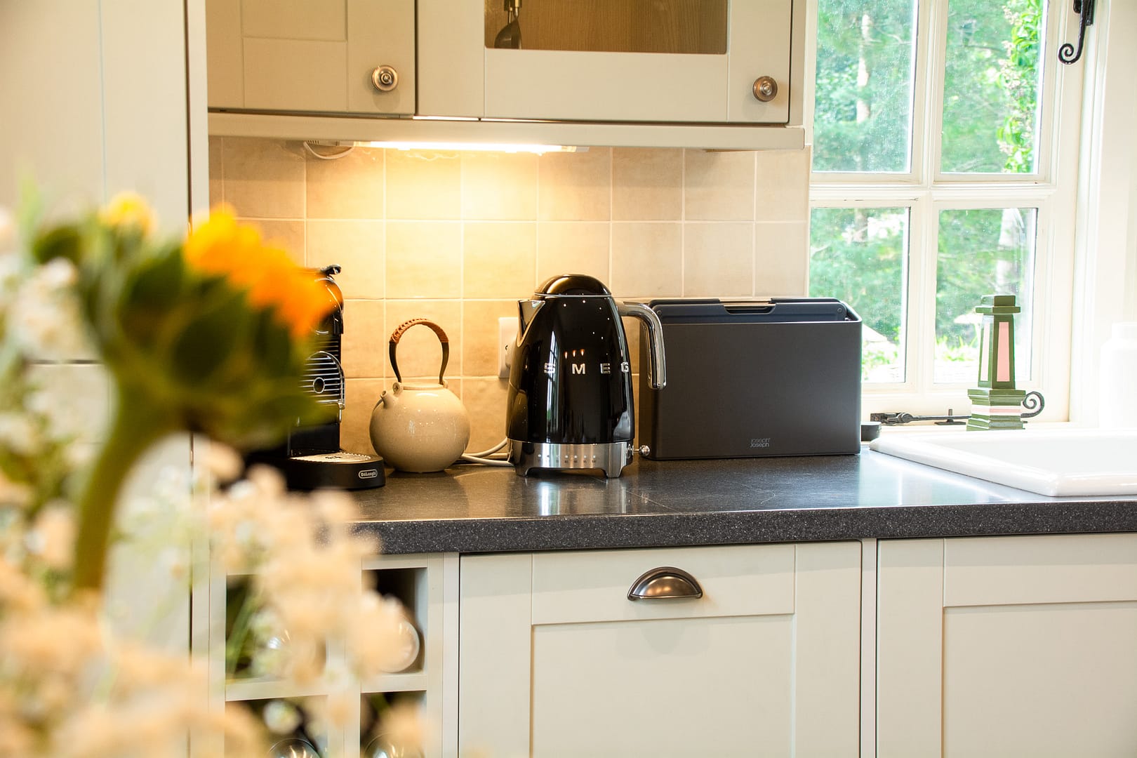 Light kitchen units with black worktops with a black smeg kettle and chopping boards near a window.