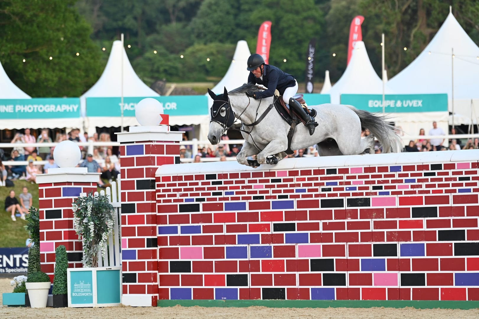 An equestrian rider skilfully jumps over a decorative puissance fence at the Bolesworth International, with the Bolesworth trade stands visible in the background.