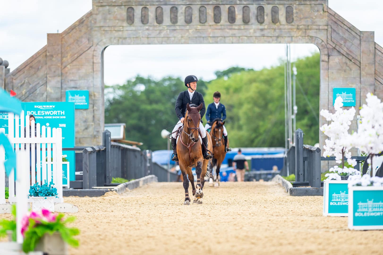 Two equestrian riders on horseback enter the arena at the Bolesworth International Horse Show, framed by the iconic stone archway with event branding and floral decorations.