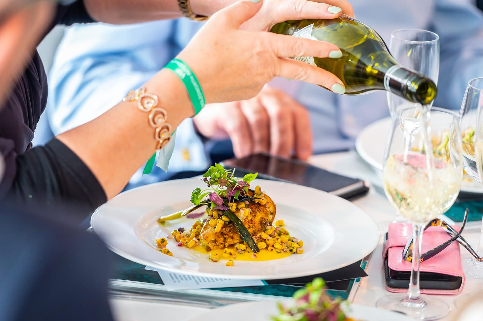 A close-up of a gourmet plated dish topped with greens, as someone pours white wine into a glass at an elegant dining setting.
