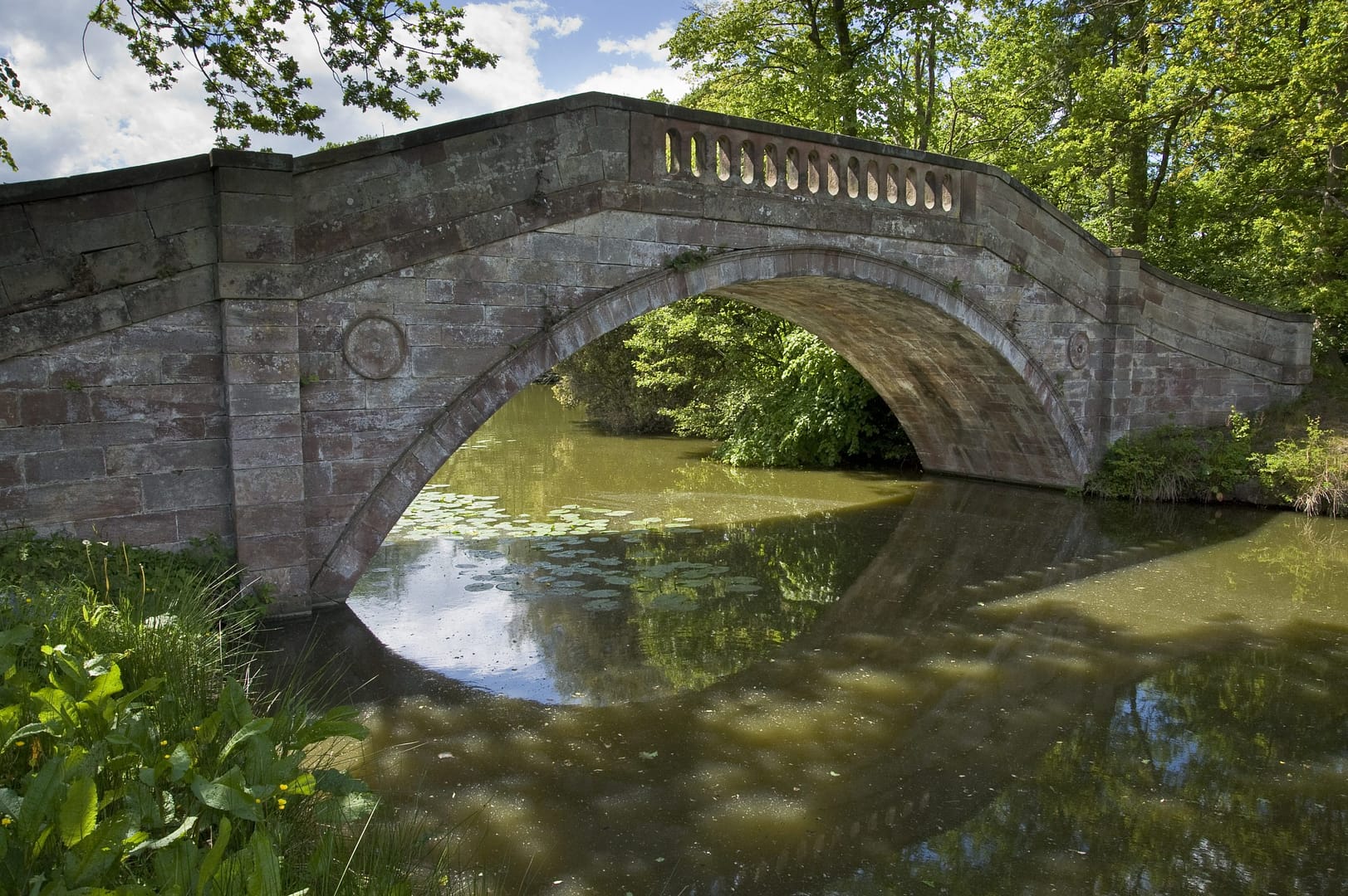 A stunning historic stone bridge arching over a tranquil lake at Bolesworth, Cheshire. The bridge is surrounded by lush greenery, with reflections shimmering in the water and lily pads floating below.