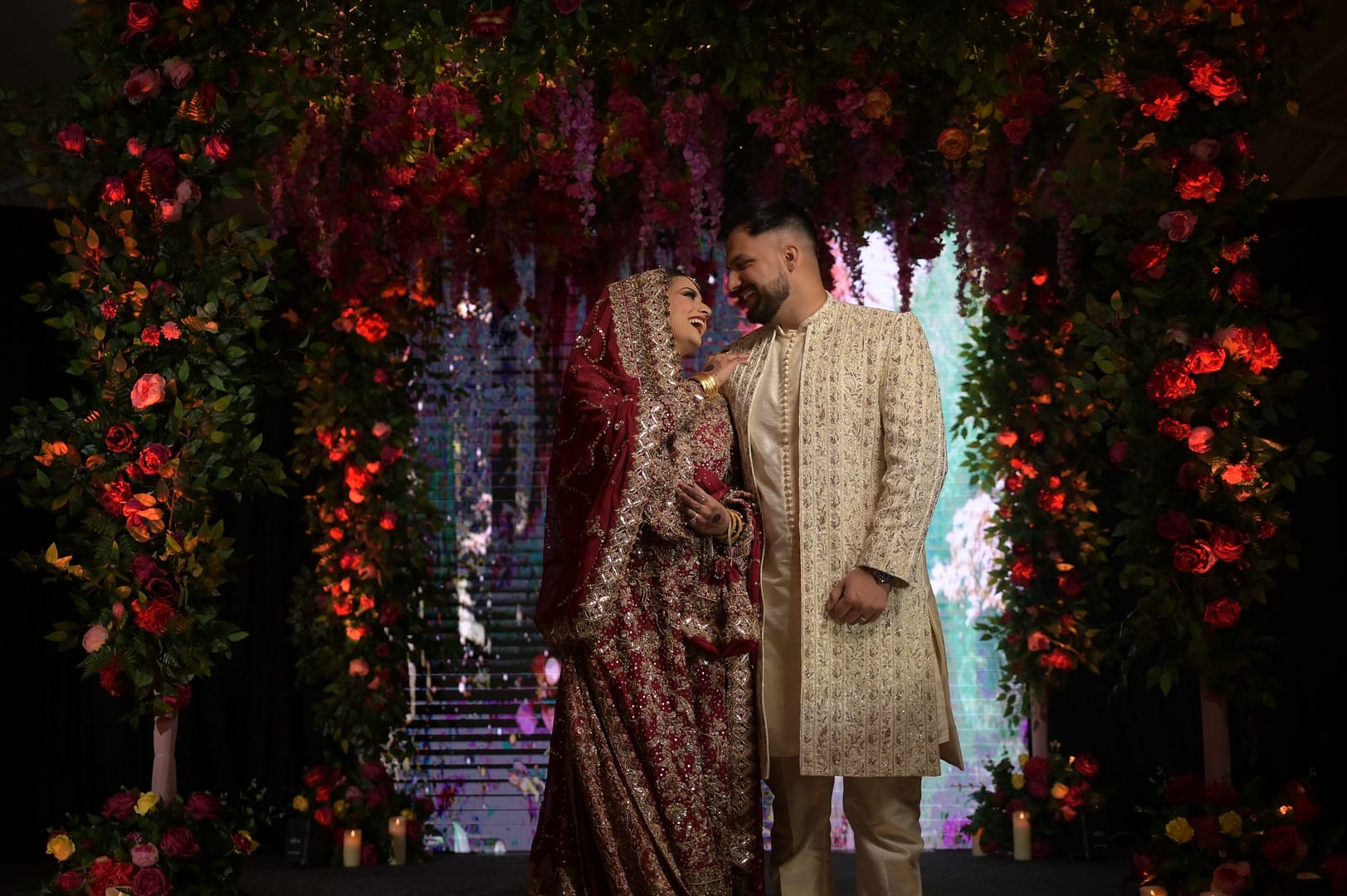A newlywed South Asian couple shares a joyful moment under a breathtaking floral archway at their wedding reception. The bride dazzles in a deep red and gold bridal lehenga with intricate embroidery, complemented by a statement jewelry set and matching veil, while the groom looks regal in an ivory sherwani with gold embellishments. The romantic setting is enhanced by cascading floral arrangements in rich red, pink, and orange hues, with warm ambient lighting creating a magical, intimate atmosphere.