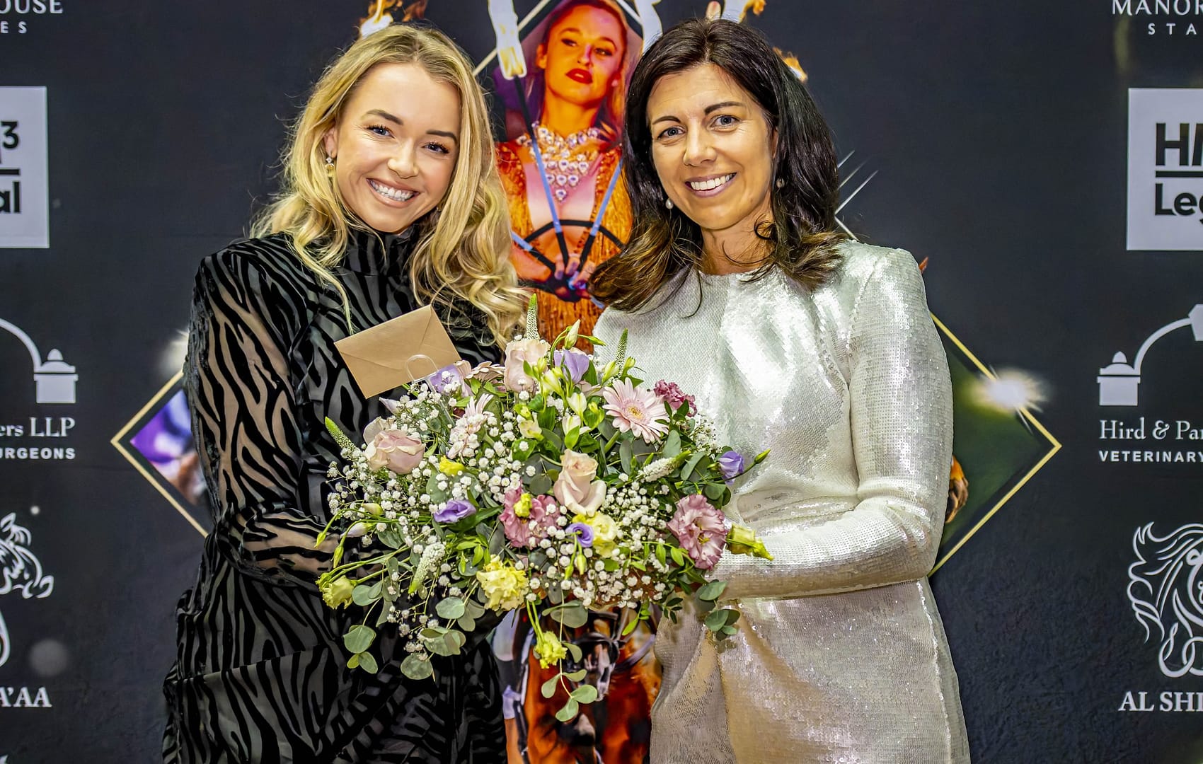 Two elegantly dressed women smiling and holding a bouquet of flowers at the Bolesworth Elite Auction 2024. The backdrop features luxury brand logos and a striking event design, highlighting the prestigious equestrian auction atmosphere.