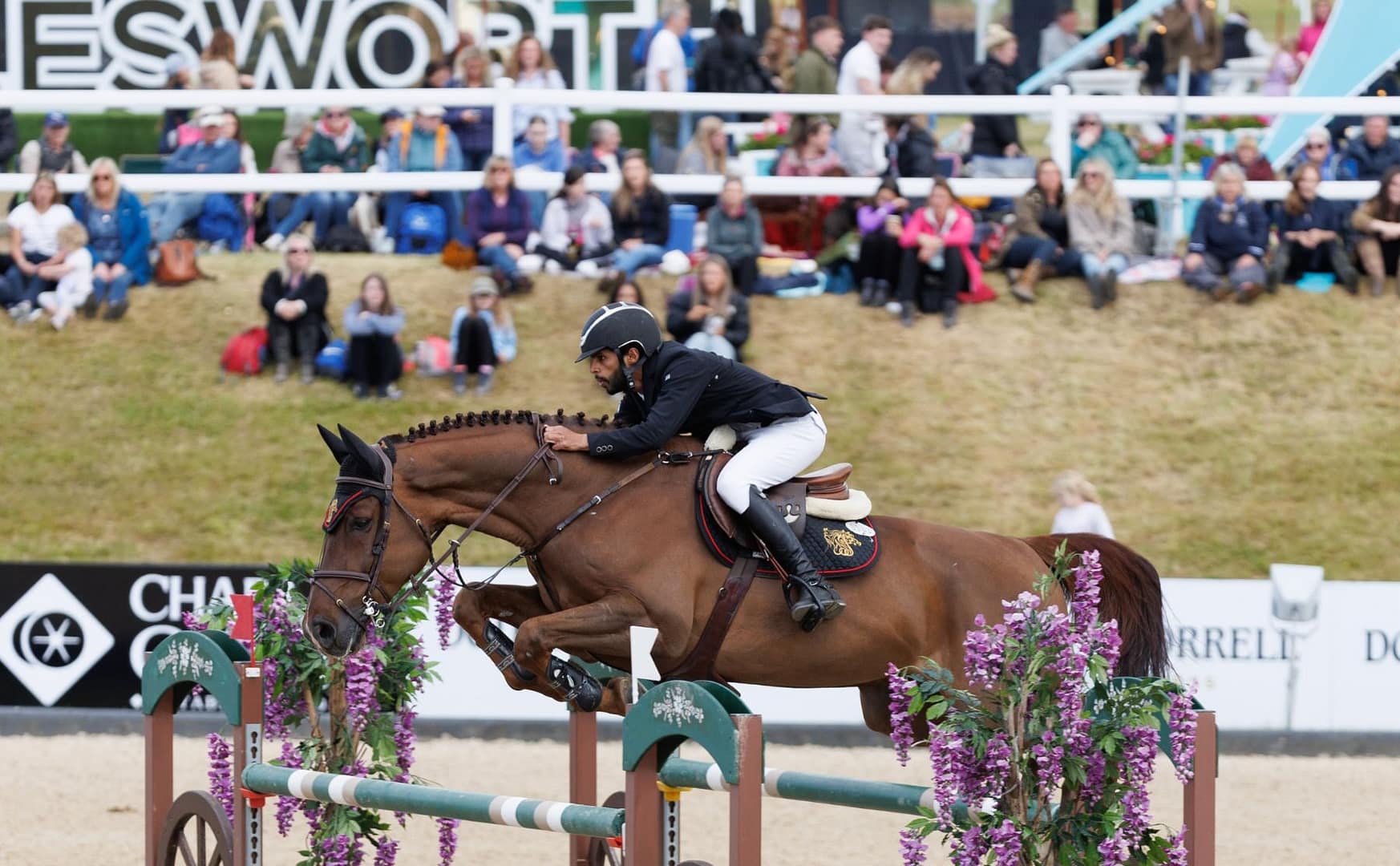 Al Shira'aa showjumping rider clears a jump at Bolesworth International Horse Show, with a focused crowd watching in the background.