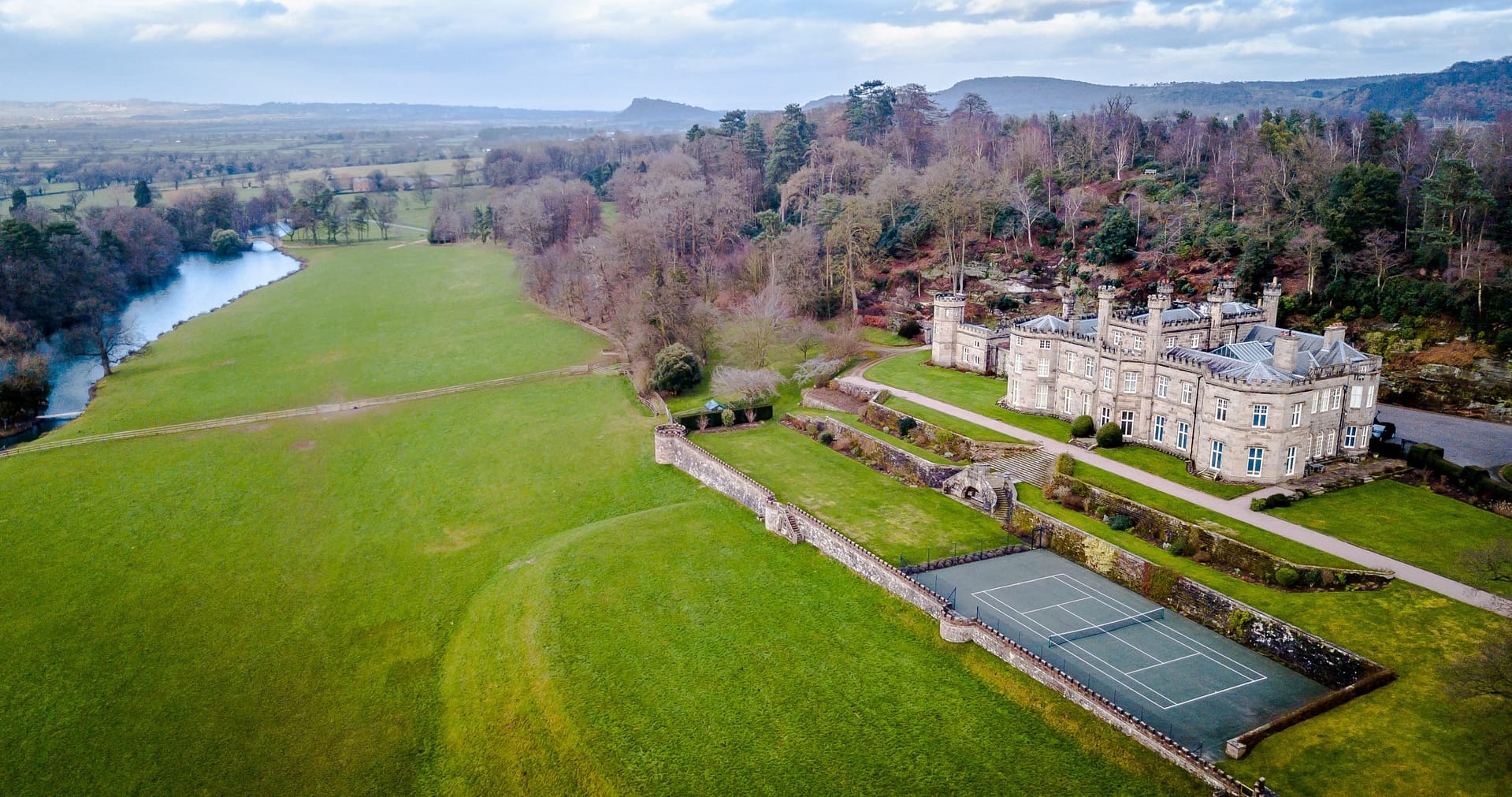 A stunning aerial view of Bolesworth Castle, showcasing its historic architecture, landscaped gardens, and scenic countryside, with a tennis court and lake visible in the estate.