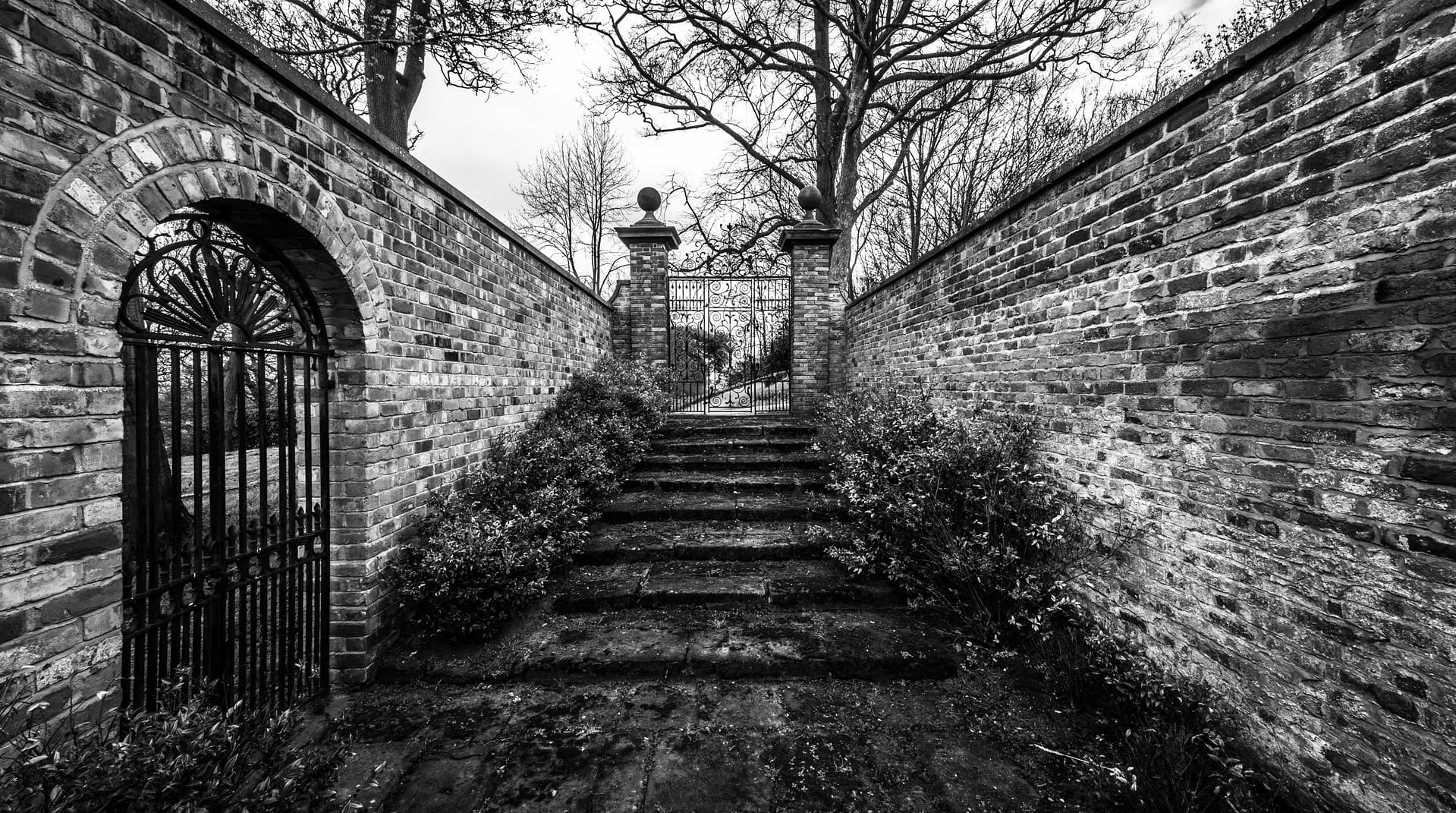 A black and white photograph of a historic brick-walled pathway and ornate wrought-iron gate at Bolesworth Estate. The weathered stone steps, aged brick walls, and intricate gate design evoke a timeless, mysterious atmosphere, leading visitors towards a hidden section of the estate.