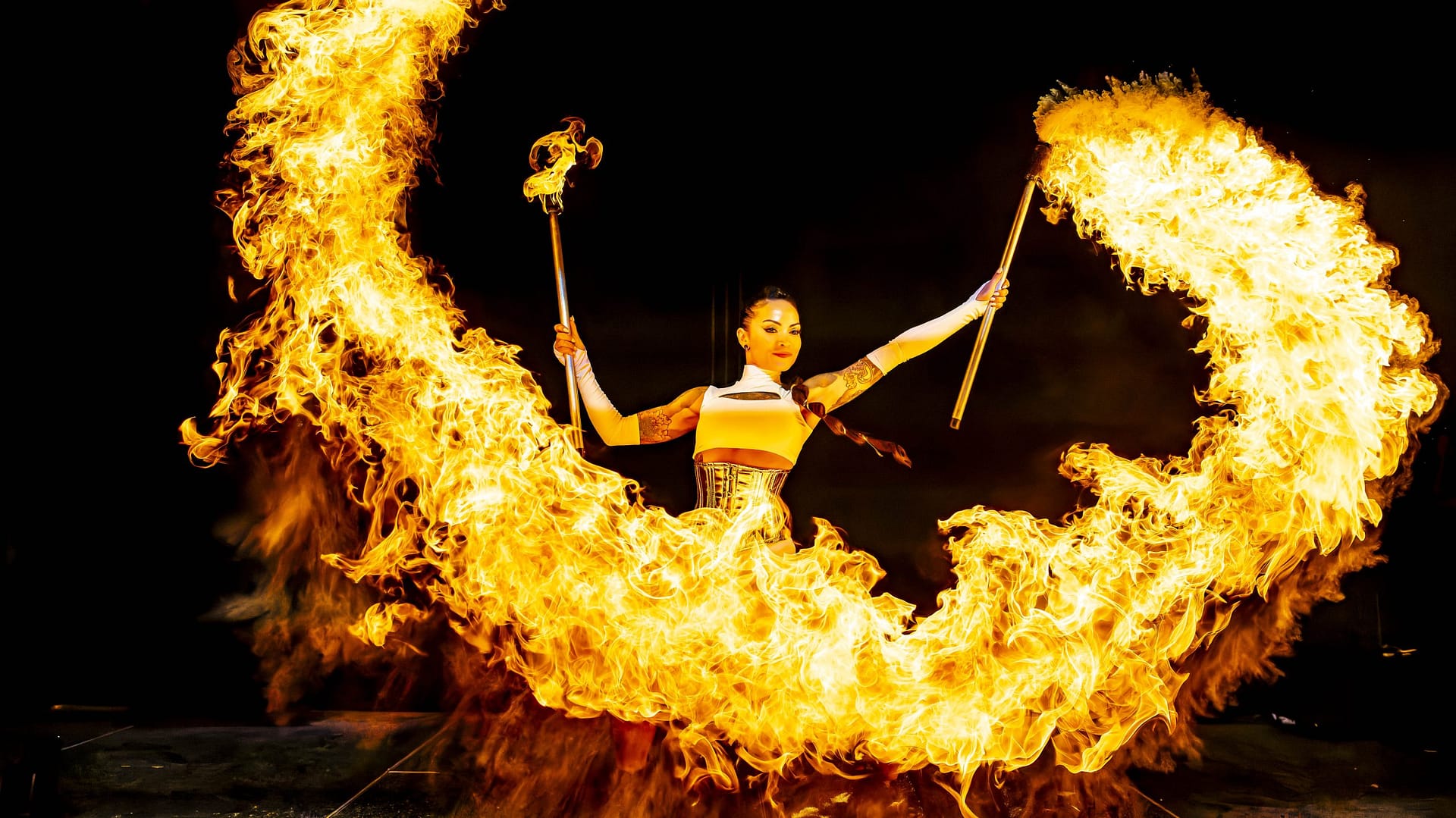A daring fire performer wields flaming torches, creating a mesmerizing arc of fire at the Bolesworth Elite Auction. Dressed in a gold corset and white bodysuit, she exudes confidence and energy while showcasing her spectacular fire skills against a dark, dramatic backdrop.