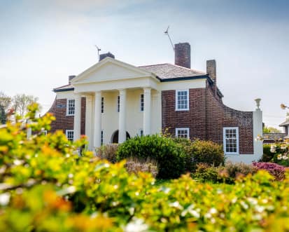 A house with classical columns, surrounded by greenery under a partly cloudy sky