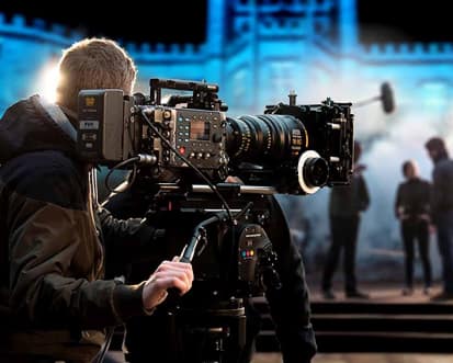 A film crew operating a professional camera during an outdoor event with Bolesworth Castle lit-up in the background