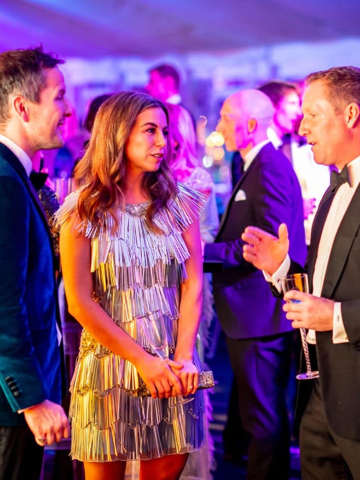 Elegant guests at the Bolesworth Crystal Ball, dressed in formal attire, engaging in conversation under vibrant event lighting.