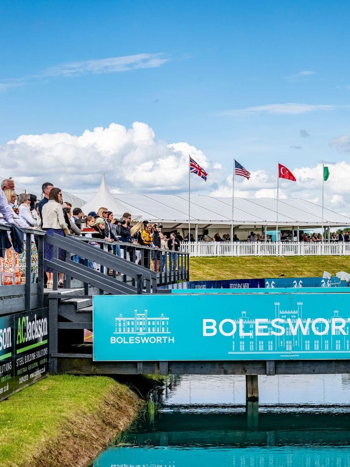 A vibrant outdoor scene at Bolesworth, featuring spectators standing on a platform overlooking the international show ground with international flags waving in the sky.