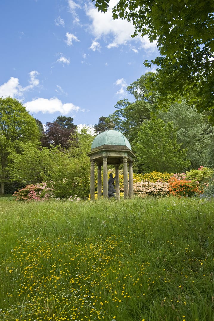 A beautifully preserved historic pavilion, containing Diana the Huntress, nestled among lush greenery and wildflowers at Bolesworth Open Gardens, Cheshire. The dome-shaped structure is surrounded by vibrant azaleas and rhododendrons, set against a bright blue sky.